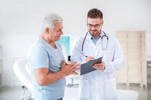 A white-haired man wearing a blue shirt is in an office receiving guidance from a doctor wearing glasses, a white coat, and holding a clipboard.
