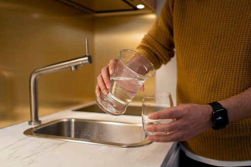 A man in a kitchen is near a sink and transfers water from a small bottle to a glass.