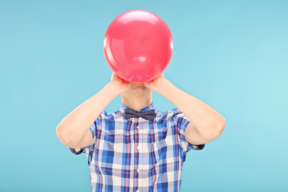 Indigo blue background with a white man wearing a checkered blue-toned shirt and a blue bow tie, inflating a balloon at face level, looking upwards, representing 'How to reduce an enlarged prostate.