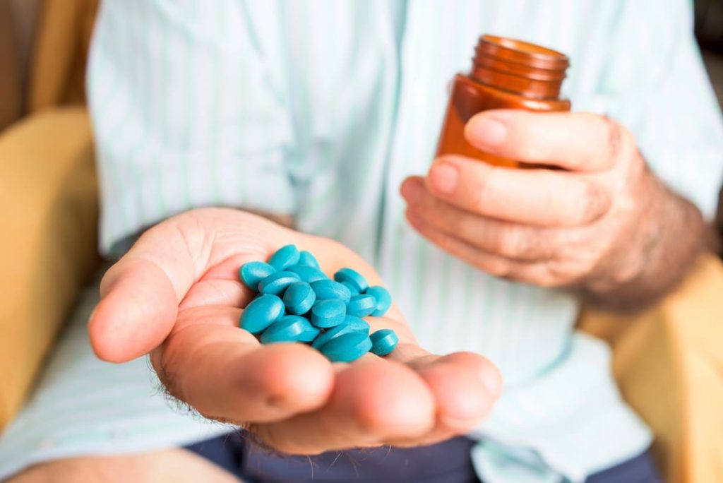 white man sitting in the background with extended arm holding blue pills while the other hand holds a medicine bottle simulating if Viagra needs a prescription