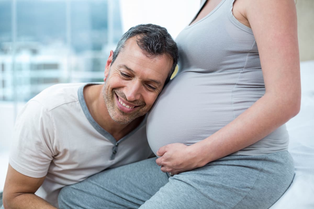 Middle-aged white man resting his head on a pregnant white woman's belly. Both are wearing grayish clothes. The image represents the concern of how to know if I am infertile