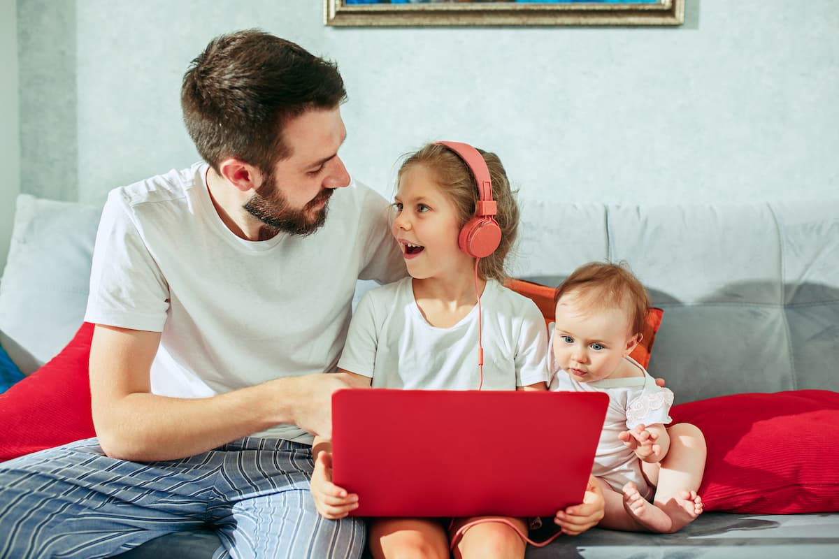 A young, white father with a white t-shirt, jeans, beard, and brown hair sits on the couch looking at his two children, a white girl with a white t-shirt, orange headphones, and a red notebook on her lap, sitting on the couch, and a white baby wearing white clothes. The image represents how to know if I am infertile.