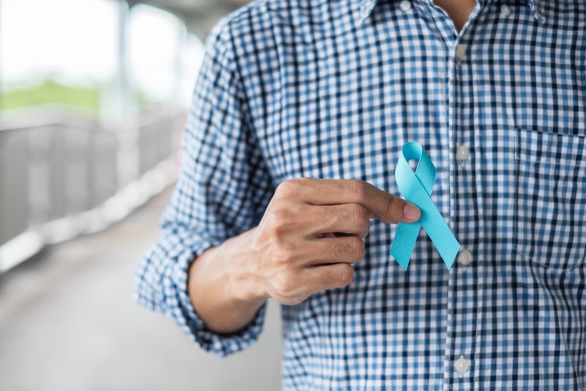 Chest of a man wearing a blue and white checkered shirt holding a blue ribbon representing prostate cancer myths and facts in a blurred background