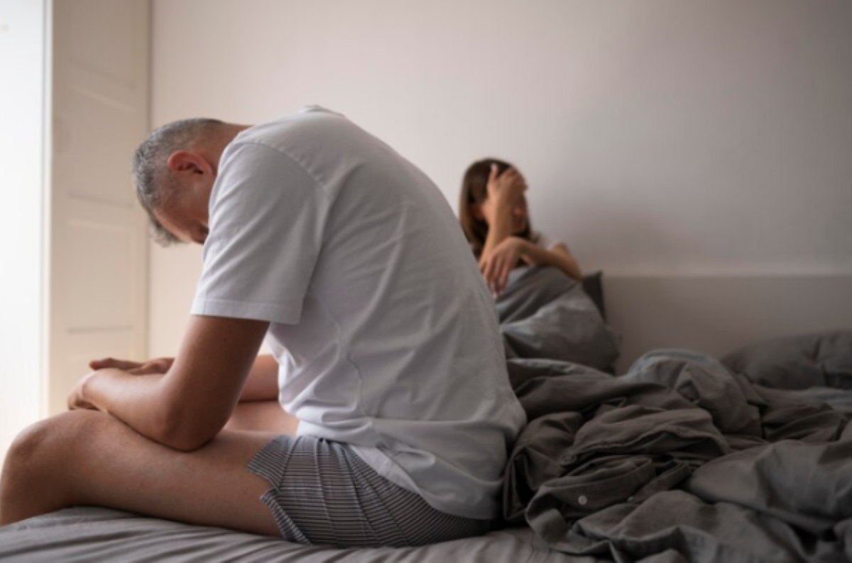 Homem cabisbaixo sentado na beira da cama. Uma mulher está ao fundo com a mão no rosto. O casal age como se o homem sofresse com disfunção erétil