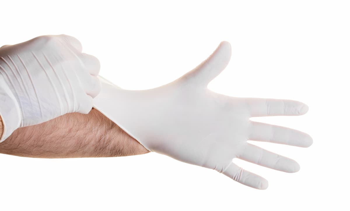 Mão de homem branco vestindo uma luva médica descartável de látex, puxando-a para ajustar em sua mão. A cena sugere a preparação para um exame para avaliar qual é a função da próstata