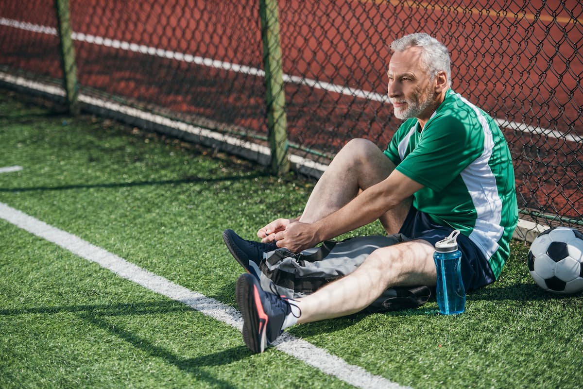 Homem branco grisalho idoso se preparando para partida de futebol do Projeto PCaGOAL sentado no gramado verde encostado em alambrado colocando o tênis