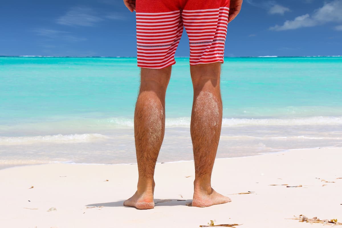 A man’s legs on the beach wearing striped red and white swim trunks, standing on light sand, illustrating if heat increases penis size
