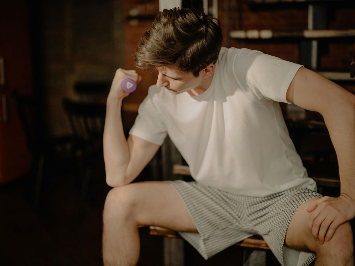 man sitting wearing loose cotton boxer shorts and a white t-shirt, holding a dumbbell