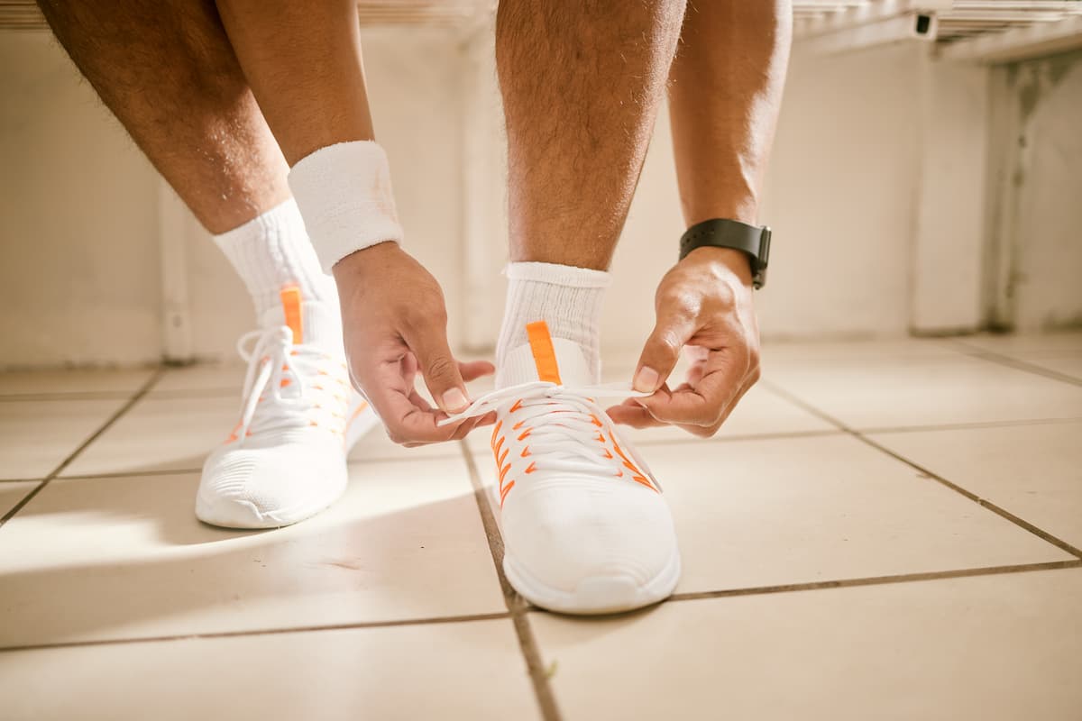 White tiled floor, man standing, focus on feet with white sneakers and hands tying shoelaces representing an exercise to avoid erectile dysfunction