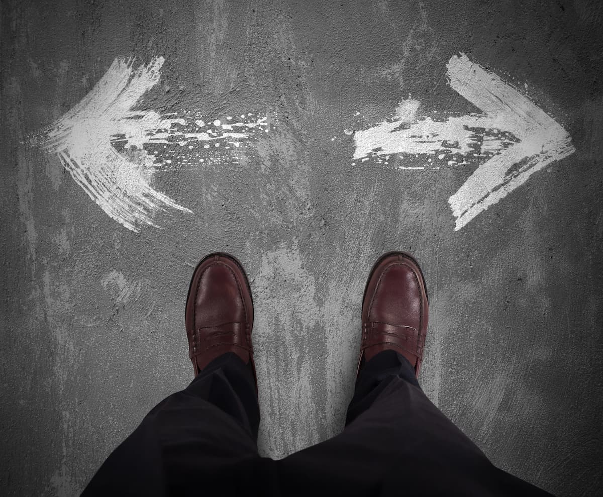 Pés com sapatos marrons em frente a duas setas brancas pintadas no chão, uma apontando para a esquerda e outra para a direita, sugerindo um dilema quanto a prótese peniana ideal segundo Dr. Paulo Egydio. O fundo é uma superfície cinza com aparência desgastada