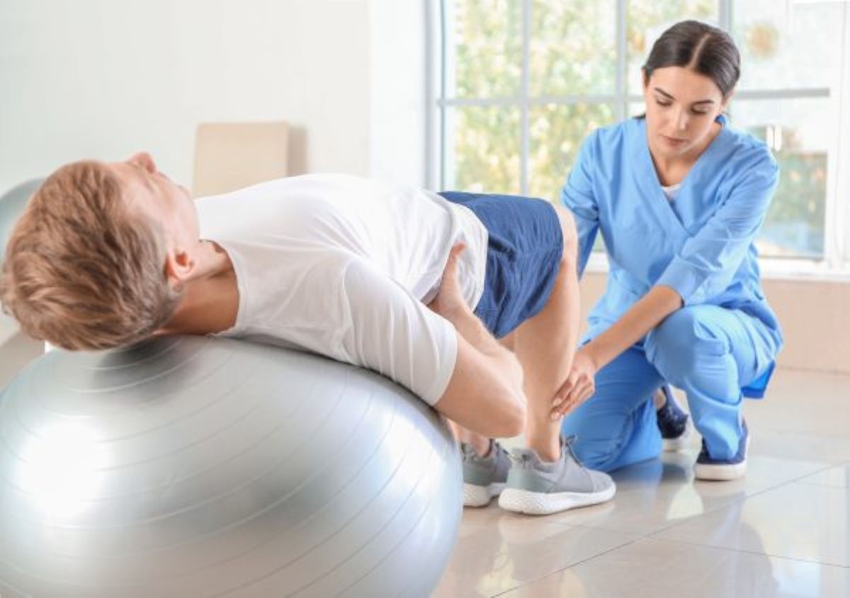 Homem branco de camiseta branca e bermuda azul deitado sobre uma bola de exercício enquanto uma profissional mulher em uniforme azul apoia suas pernas durante o movimento na fisioterapia pélvica.