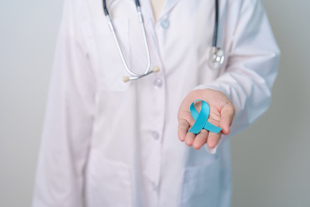 Doctor in a white coat with a stethoscope around his neck holding a blue ribbon in his hand, symbolizing myths and facts about the prostate exam.