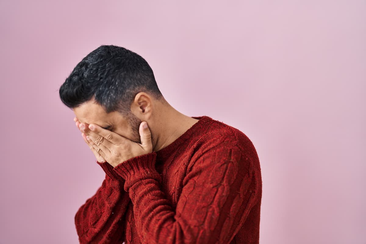 Fundo em tom de rosa claro e, em destaque, homem de perfil com cabelos curtos, usando um suéter vermelho com detalhes trançados, que está cobrindo o rosto com as mãos, transmitindo uma possível sensação de angústia ou desconforto devido a disfunção erétil aos 40 anos.