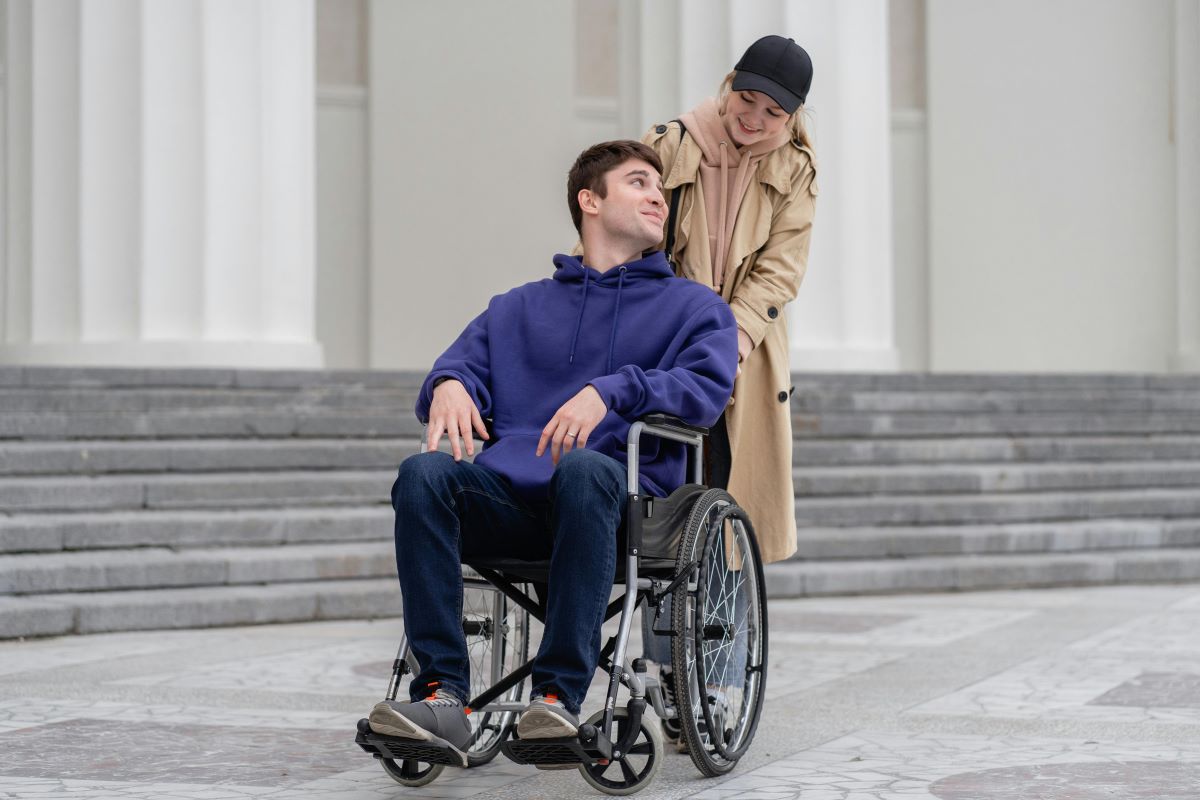 Man in jeans and a blue sweatshirt being pushed in a wheelchair by a woman in a beige coat and white cap.