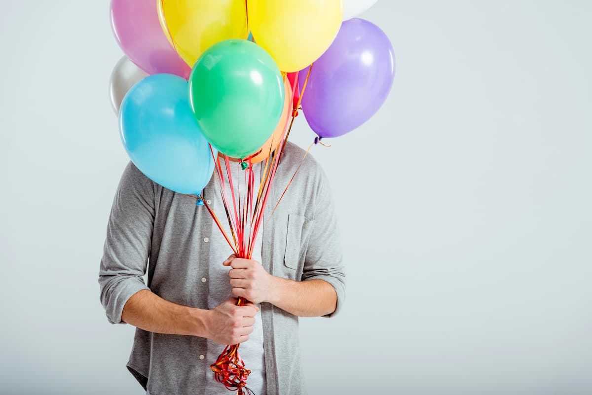 Homem de camisa cinza segurando um buquê de balões nas cores verde, azul, roxo, amarelo, etc com a face escondida pelos balões representando a saúde e os exames check-up para homem 50 anos
