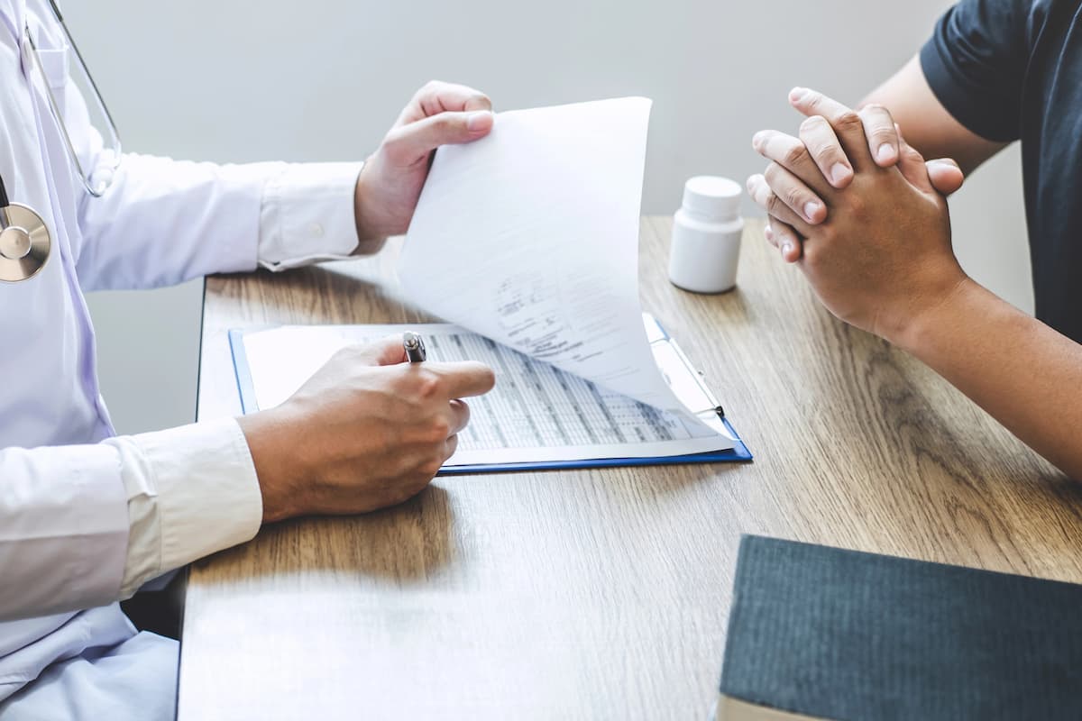 Médico vestindo jaleco branco e estetoscópio revisando um documento em uma prancheta, enquanto conversa com um paciente sentado à frente. O paciente, de camiseta preta, mantém as mãos entrelaçadas em postura de escuta. Sobre a mesa de madeira, há também um frasco de medicamentos branco e um livro, no consultório médico para detectar quais são os primeiros sintomas de problemas na próstata