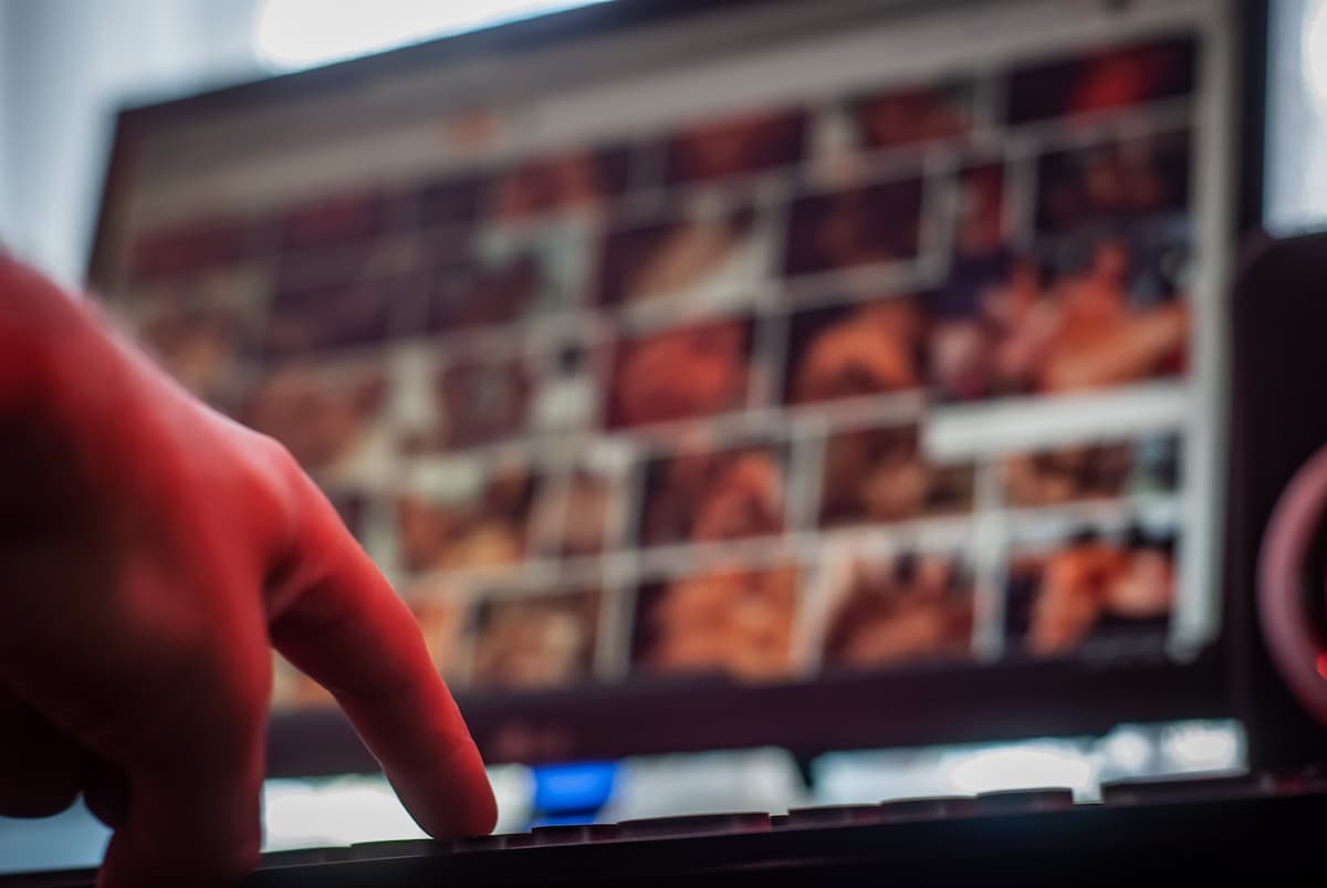 Mão se aproximando de um teclado, com um monitor ao fundo exibindo várias imagens desfocadas que parecem conter conteúdos pornográfico, que é um dos sintomas do vício em sexo