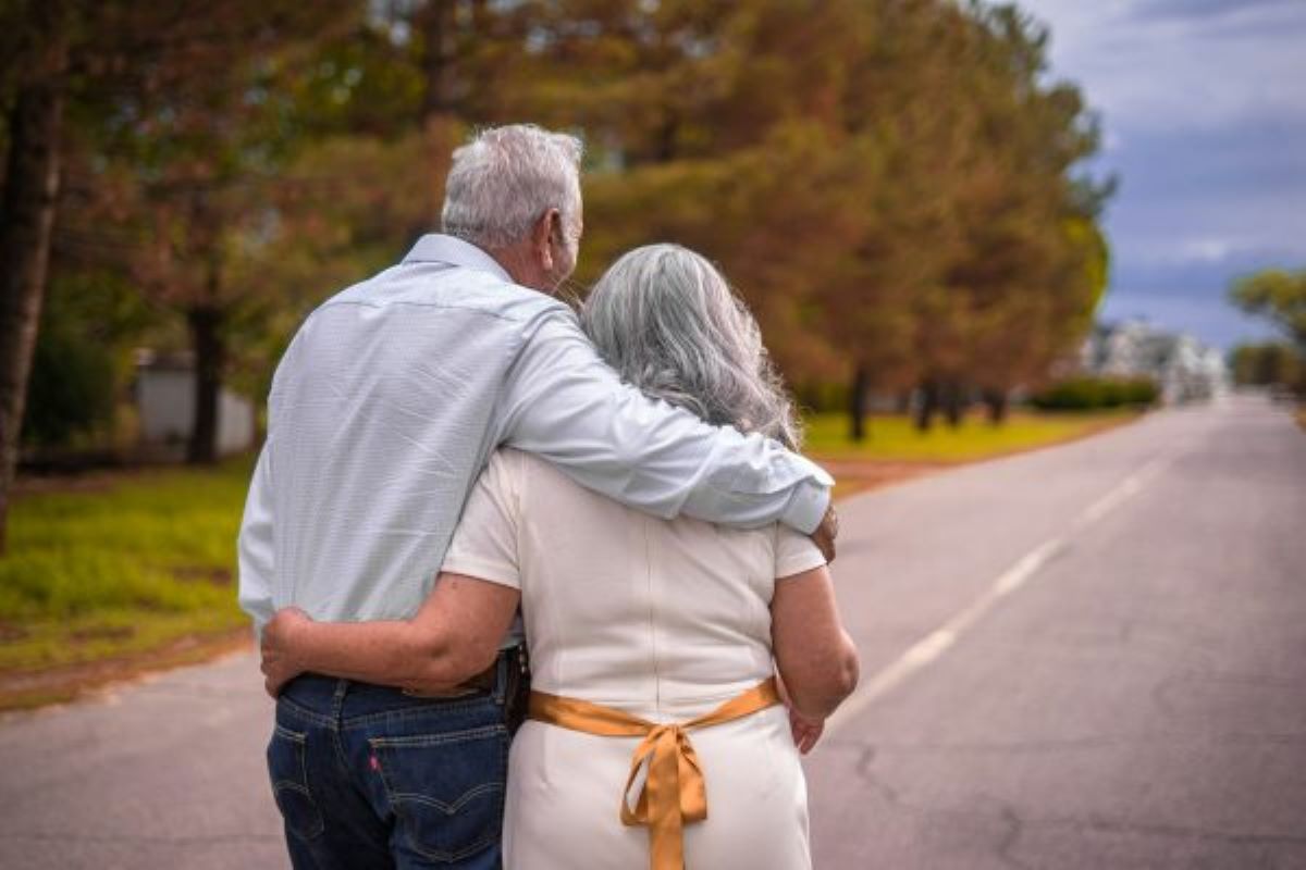 an elderly man and woman walking on a deserted street with trees. both photographed from behind