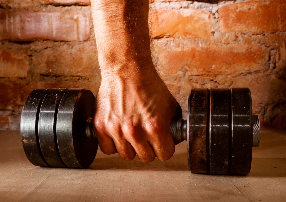 Mão firme puxando um haltere pesado, com um fundo de tijolos aparentes, representando exercícios para melhorar o desempenho sexual