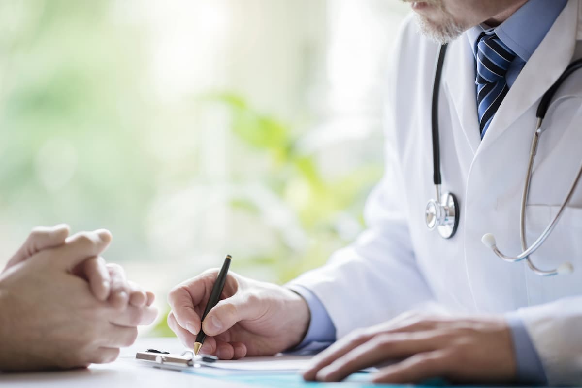 Doctor wearing a white coat and stethoscope writing notes about colorectal cancer cure on a clipboard, with a patient's hands crossed in the foreground