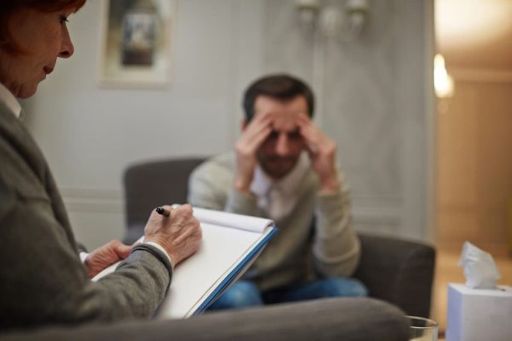 Un hombre preocupado con las manos en la cara habla con una mujer posicionada al lado que está tomando notas.