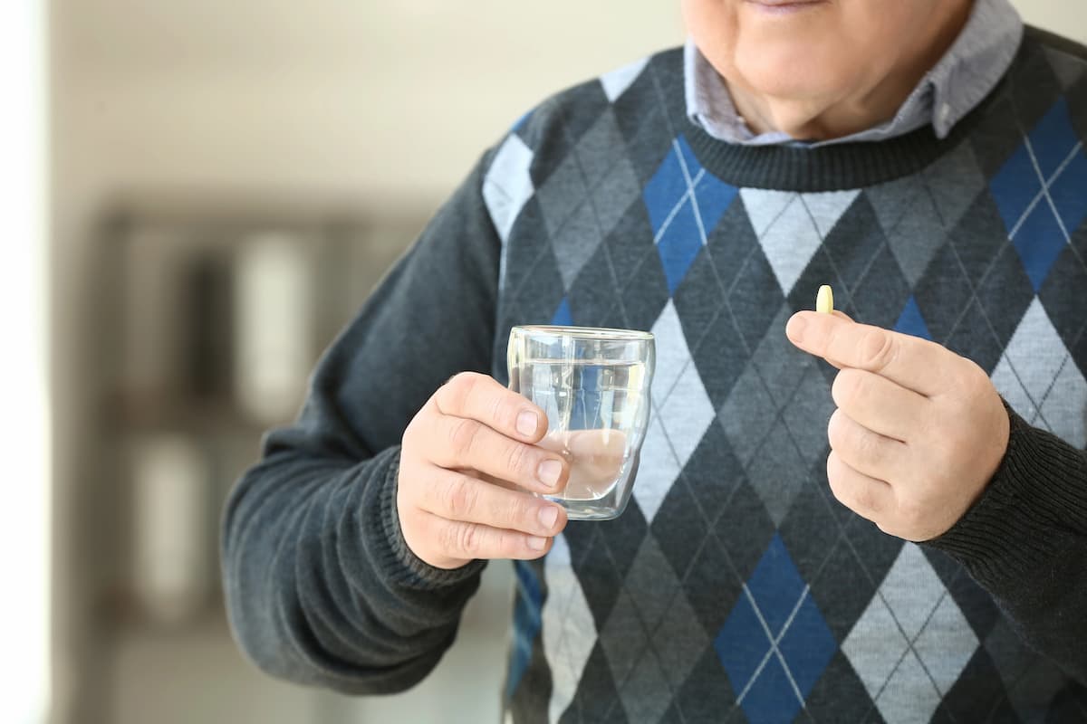 An older white man in a blue checked sweater holds a glass of water in one hand and a yellow pill in the other, illustrating whether tadalafil increases size