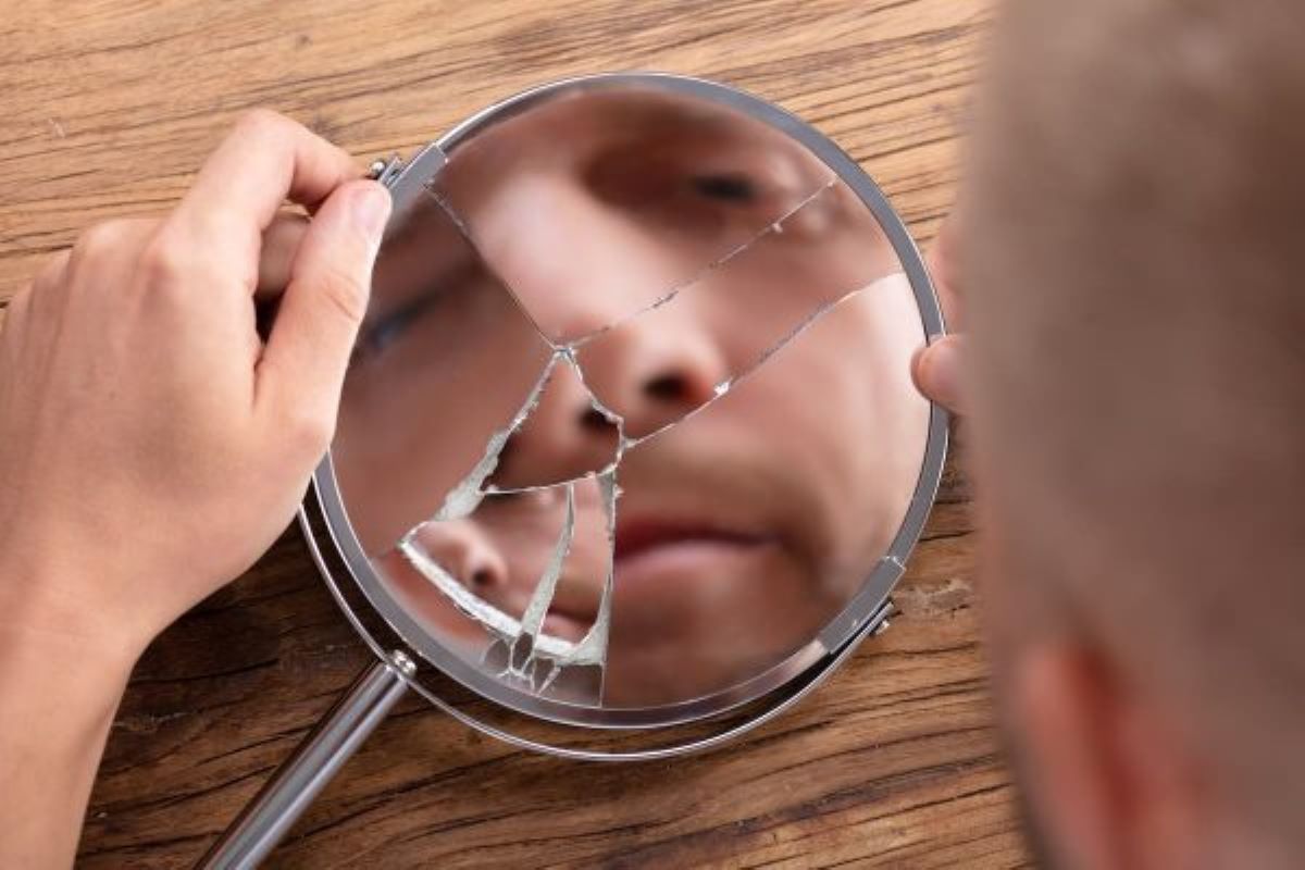A white man holding a broken hand mirror, reflecting his face in a fragmented way, on a wooden background, representing negative impacts on self-esteem and sexuality.