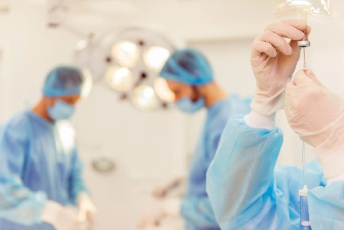 Sala cirúrgica mostra dois médicos com roupas cirúrgicas azul ao fundo, lâmpada e à frente braço e mão de médico gotejando líquido em seringa representando preparativos para uma cirurgia