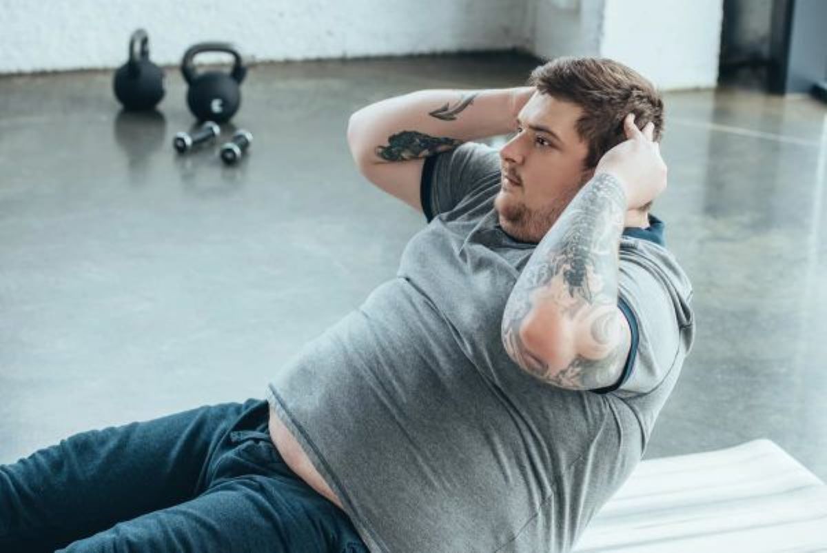 An obese white man in a gray T-shirt and blue pants doing sit-ups at the gym with dumbbells.