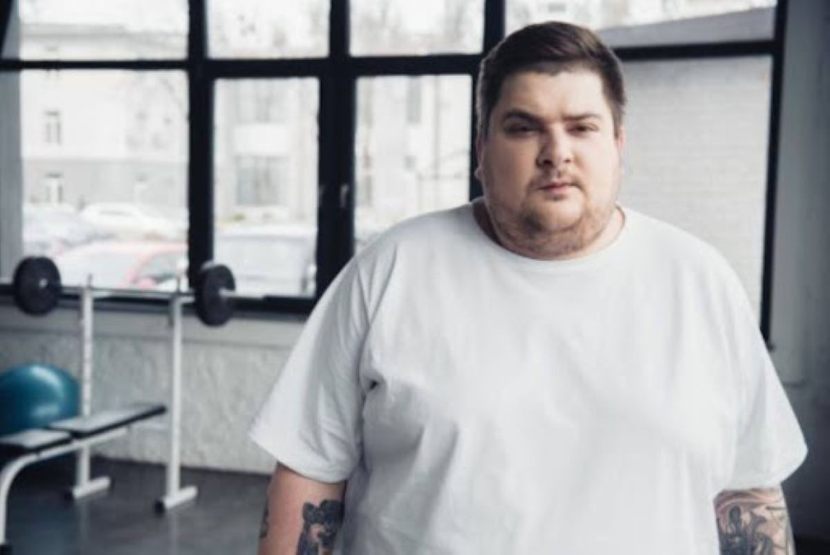 Homem branco, acima do peso usando camisa branca, tem um cenário com alguns aparelhos de ginástica.