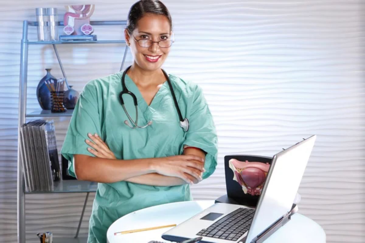 mulher branca com cabelos escuros presos, com óculos de grau, estestocópio no pescoço, roupa verde. Ela está ao clado de uma mesa redonda com um notebook sobre ela. Atrás da mulher tem uma estante e na prateleira de cima tem uma simulação do órgão reprodutor masculino.