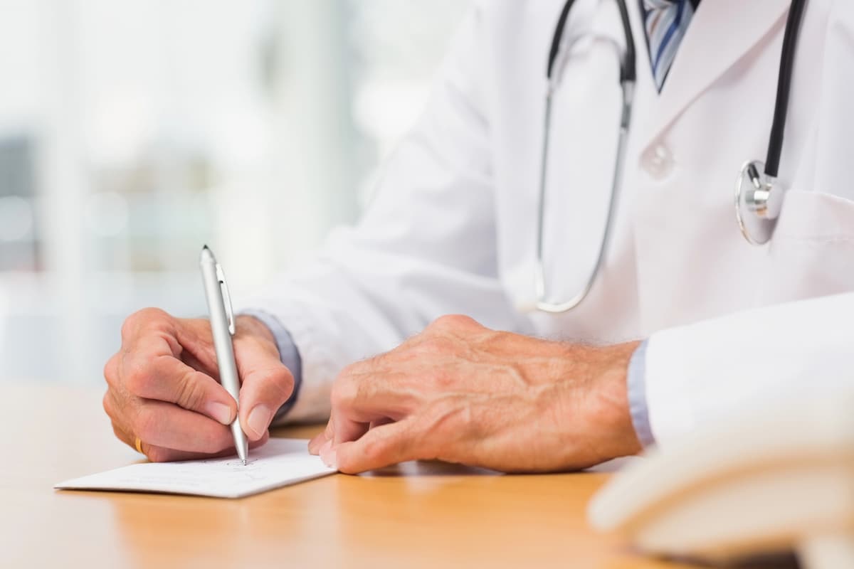 Doctor in a white lab coat writing a prescription for an erection-spray at a wooden desk, using a silver pen