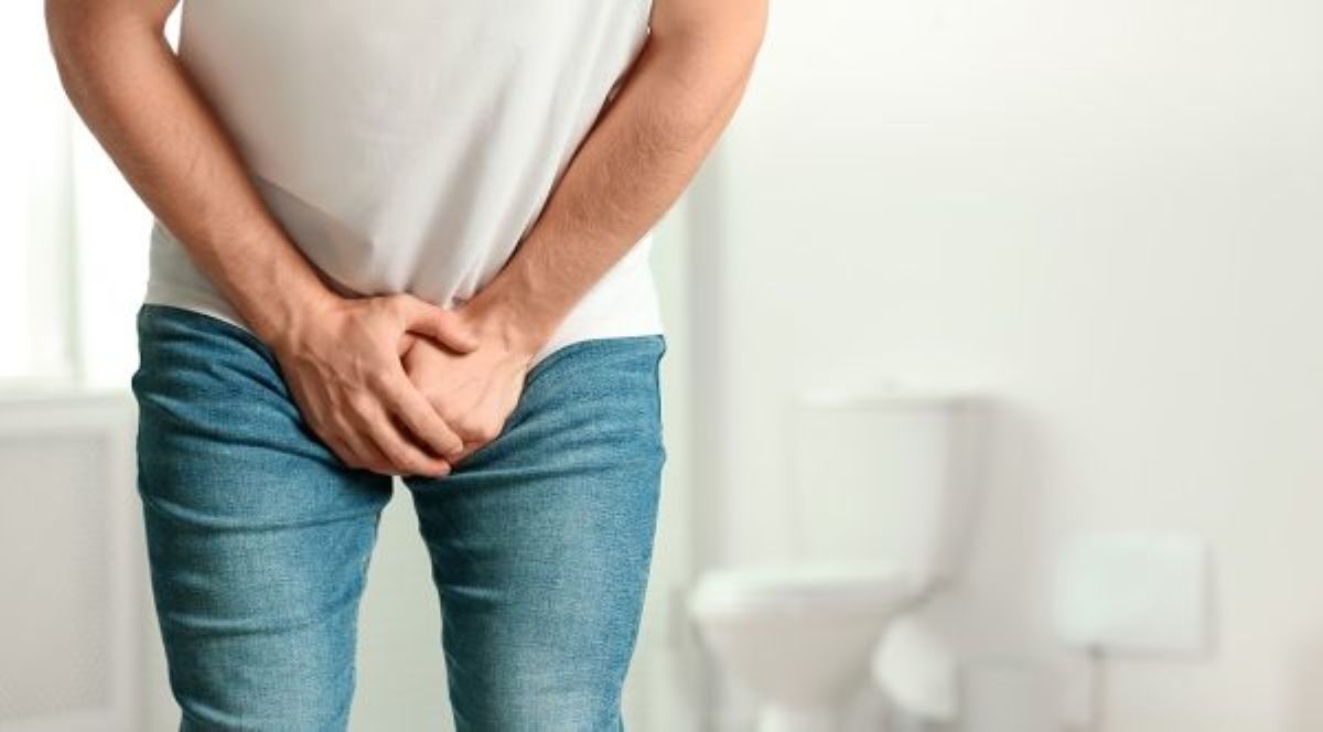 White man wearing a white shirt and jeans, hands over lower abdomen, toilet in background