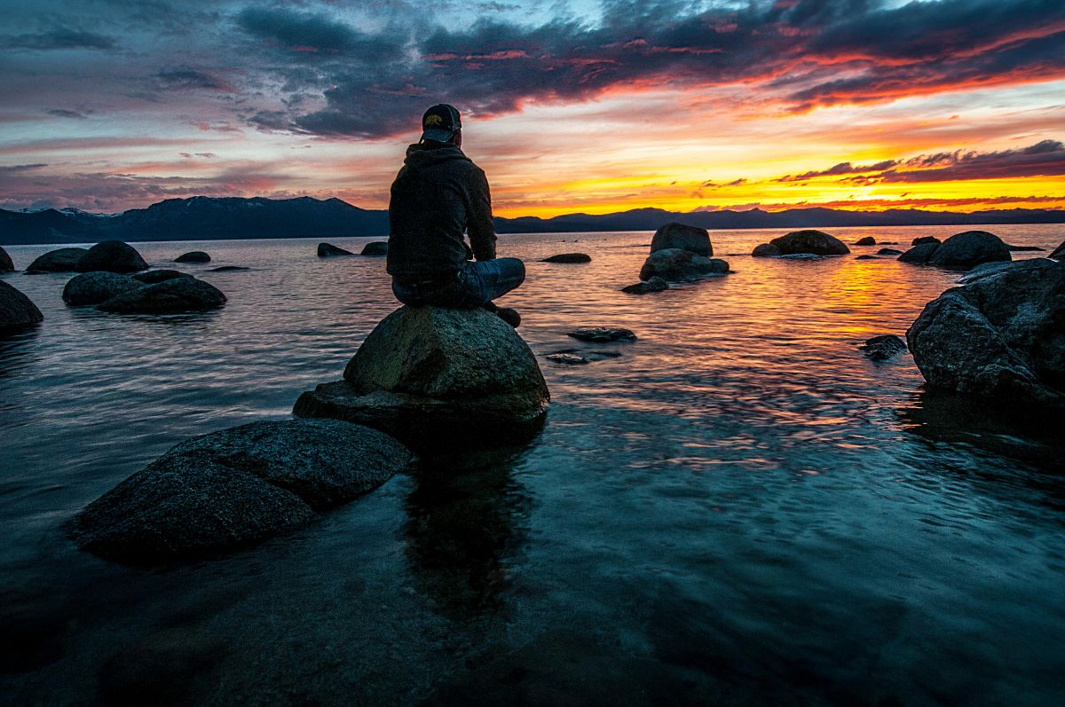 Foto que parece ter sido tirada ao final da tarde mostra um homem de boné sentado em uma pedra na parte rasa da praia. Perto dele tem outras pedras.