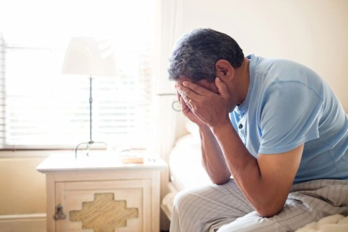 Hombre sentado al borde de la cama con las manos cubriéndose el rostro.