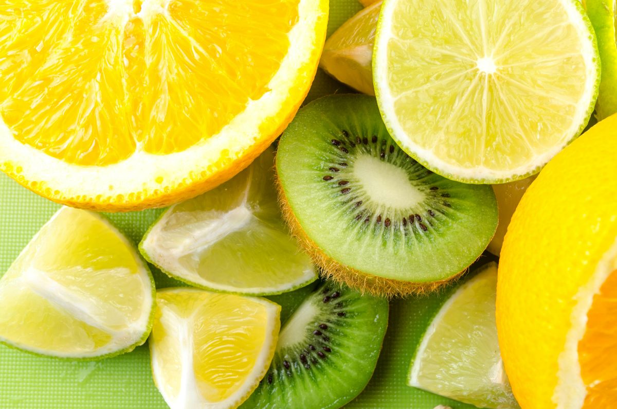 Metades de laranja, limão e kiwi, frutas ricas em vitamina C dispostas em uma superfície plana verde.