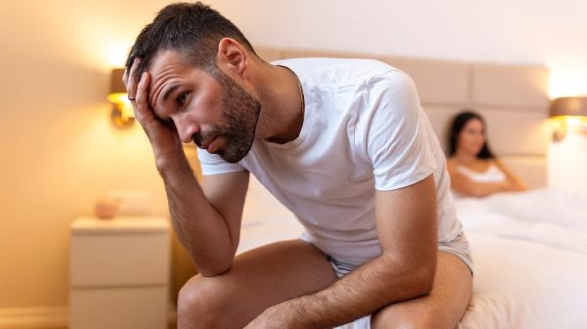Homem cabisbaixo sentado na beirada da cama. Ele veste camisa branca e short cinza, e está com a mão na cabeça, representando se mirtazapina causa impotência.