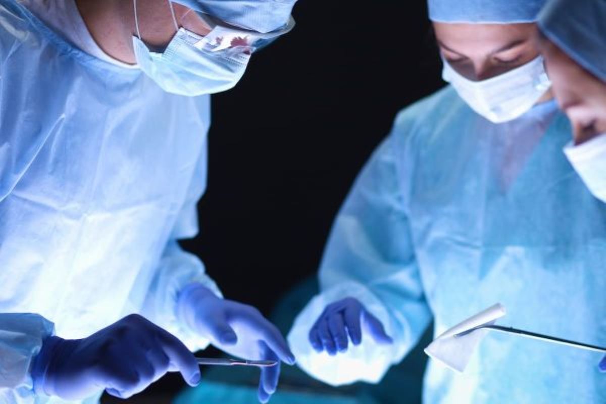 Pessoas de uma equipe cirúrgica com dois membros usando máscara, aventais e toucas azuis e luvas brancas, debruçadas durante cirurgia, representando a técnica Egydio pós-cirurgia peniana.