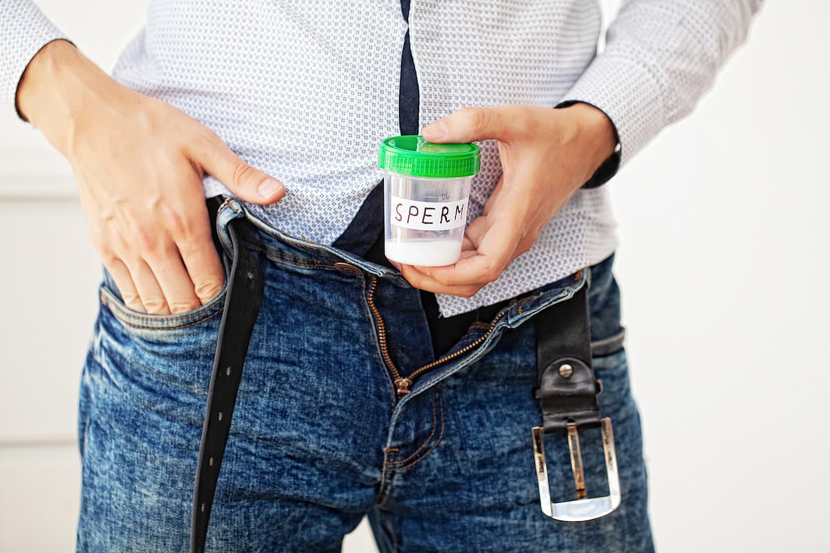 Homem branco de camisa clara e calça jeans com cinto aberto segura um frasco de exame rotulado como “esperma", simbolizando quando o homem é estéril ele ejacula.