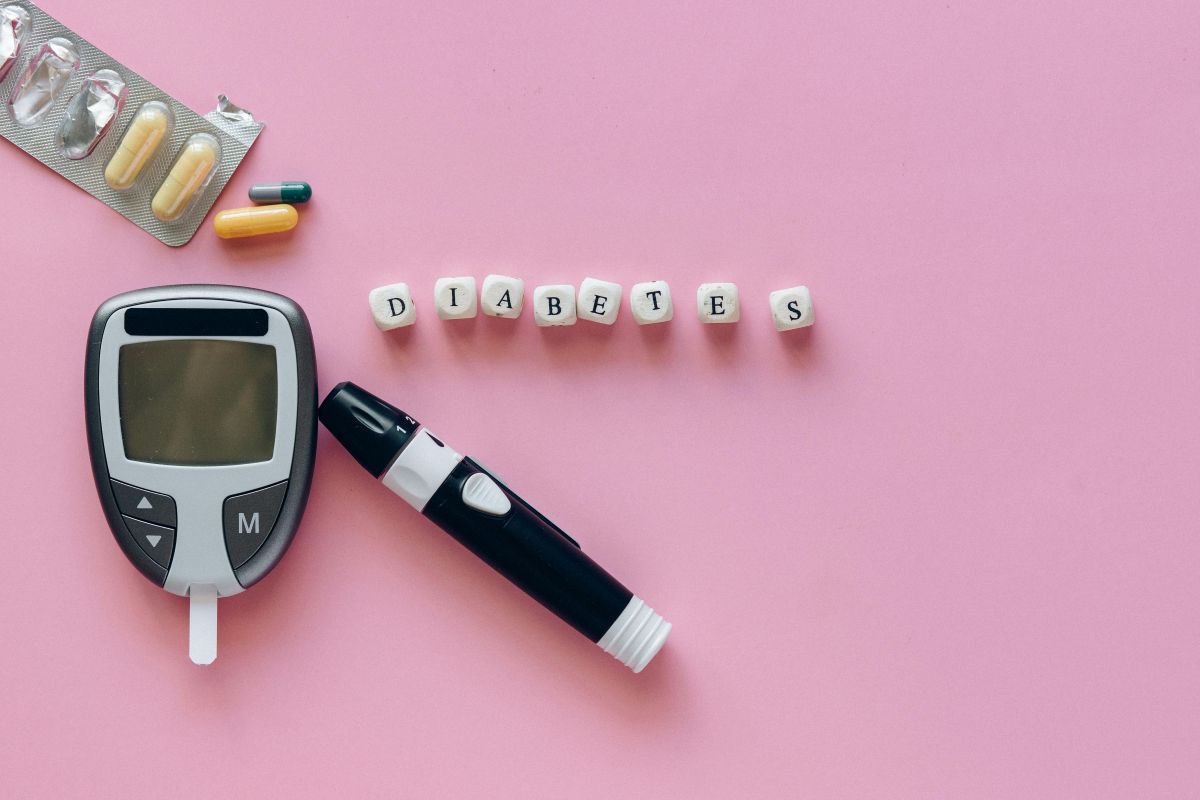 Imagem mostrando um medidor de glicose, comprimidos e a palavra 'DIABETES' escrita com blocos de letras em um fundo rosa, relacionado a importância do controle glicêmico e o tratamento do diabetes.
