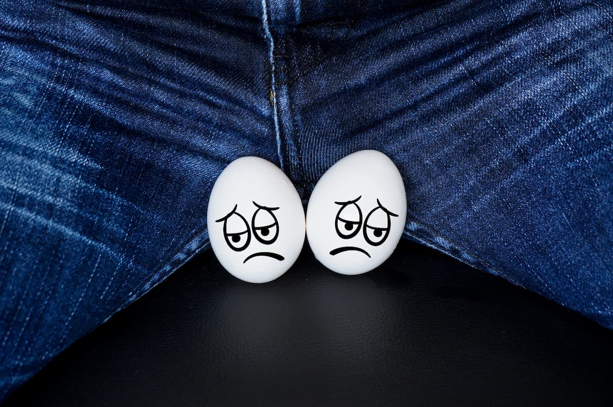 Dois ovos brancos com rostos tristes estão posicionados sobre o colo de um homem de calça jeans, simbolizando testículos e se quando o homem é estéril ele ejacula.