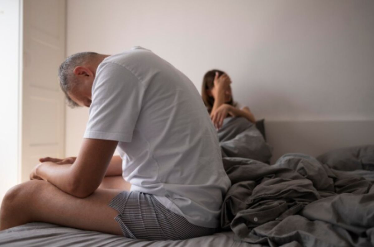 Homem usando camiseta branca e shorts cinza está sentado na cama cabisbaixo e a mulher está encostada na cabeceira da cama com ar decepcionado