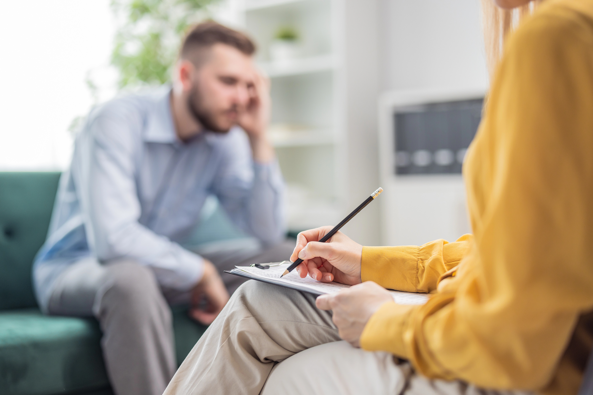 white man in neutral-toned business clothing sitting on a sofa looking reflective while a therapist in yellow writes observations on a clipboard during emotional recovery therapy after penile prosthesis surgery