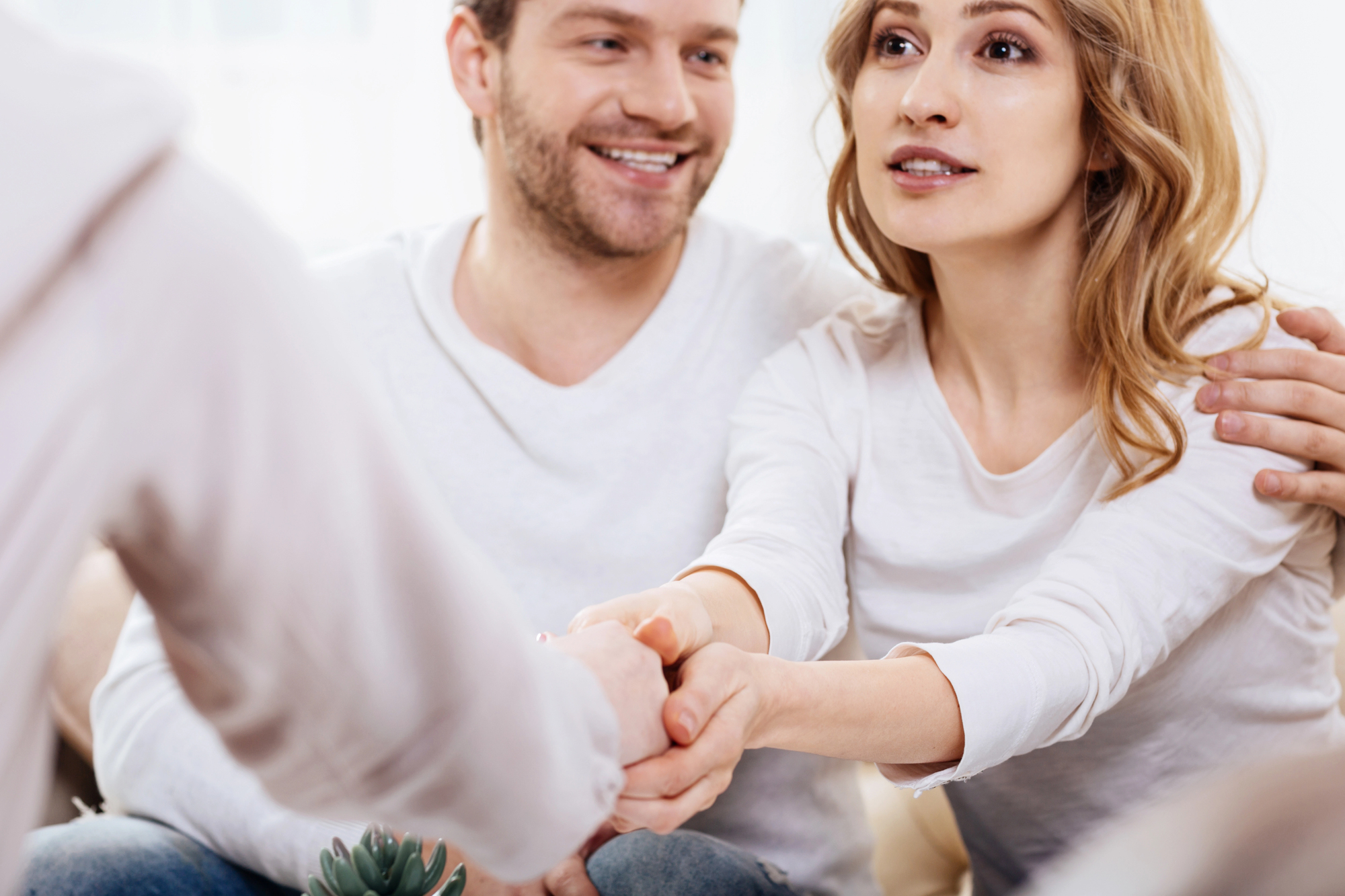 Casal branco de roupas brancas sentado, com a mulher à frente apertando a mão de um médico fora do enquadramento, conversando sobre como a parceira pode ajudar no tratamento da curvatura peniana