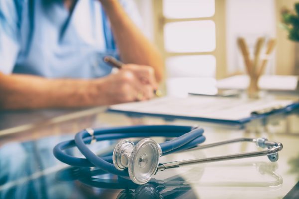Stethoscope on a glass table during a consultation about how to talk to a doctor about erectile dysfunction, with a healthcare professional in the background writing in a medical chart.