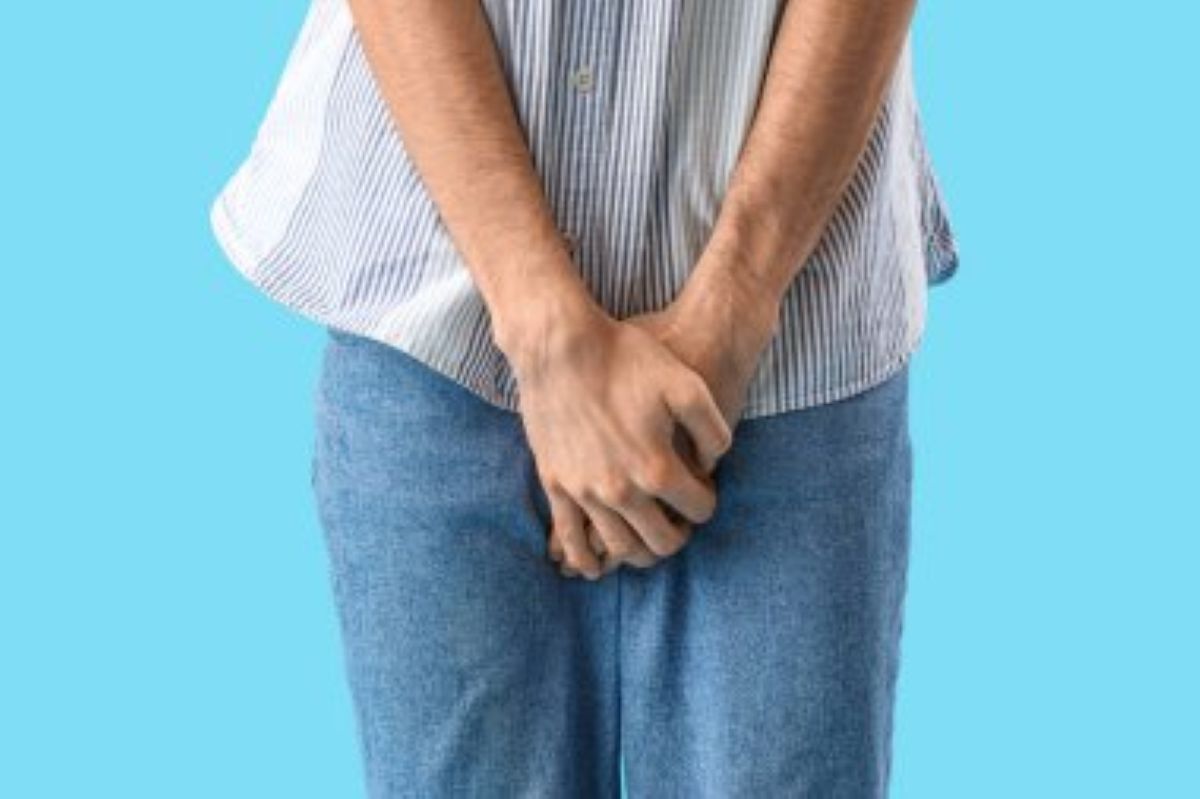 Homem usando camisa listrada e calça jeans está com as mãos sobre o pênis.