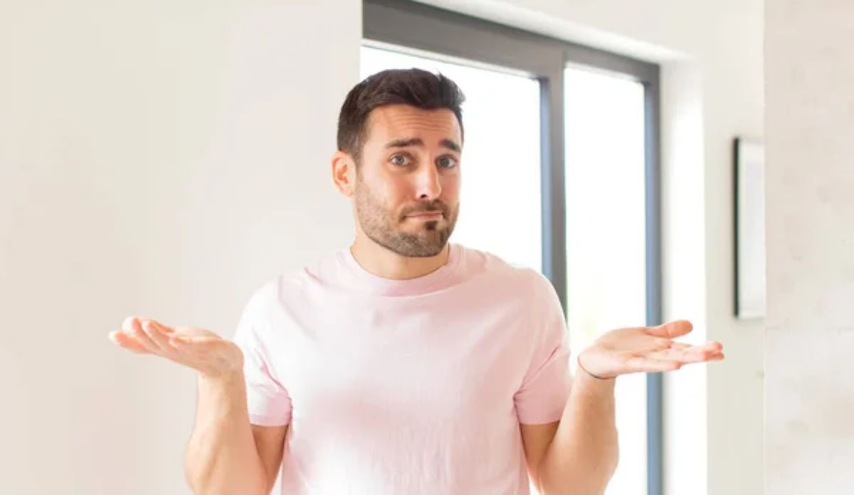 Homem branco com cabelos e barba escuros, usando camiseta branca está com os braços dobrados na altura da cintura e as mãos estão voltadas para cima. Seu semblante é de dúvida.