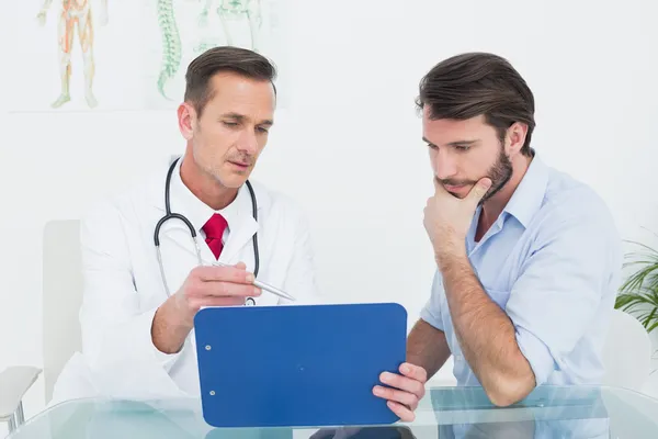 Médico usando camisa e jaleco brancos, gravata vermelha e estestocópio no pescoço segura um tablet e mostra ao paciente, um homem com camisa azul-claro com as mangas dobradas, que está com uma das mãos na boca, demonstrando atenção.