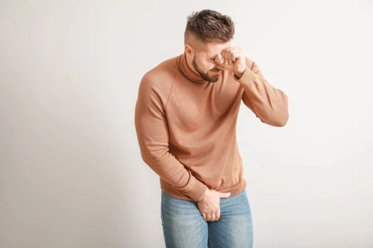 Homem usando blusa de moletom marrom e calça jeans está com uma mão sobre o pênis e a outra próxima aos olhos. Seu semblante é de dor.