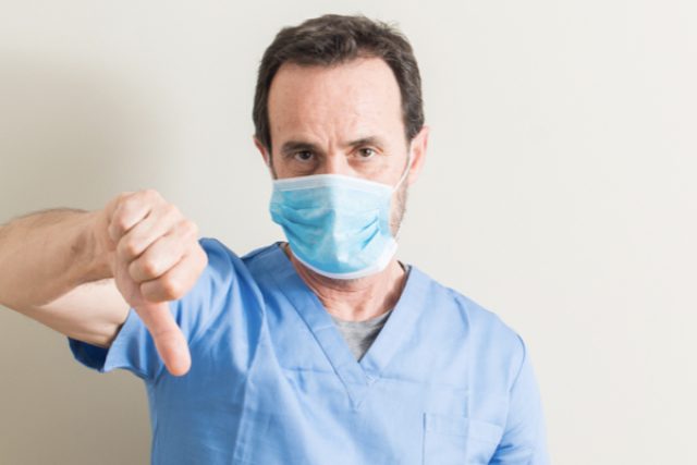 Médico usando máscara com rosto irritado, mostrando o resultado negativo da cirurgia com os polegares para baixo.